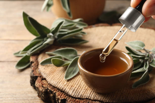Woman dropping essential sage oil into bowl at wooden table, closeup. Space for text